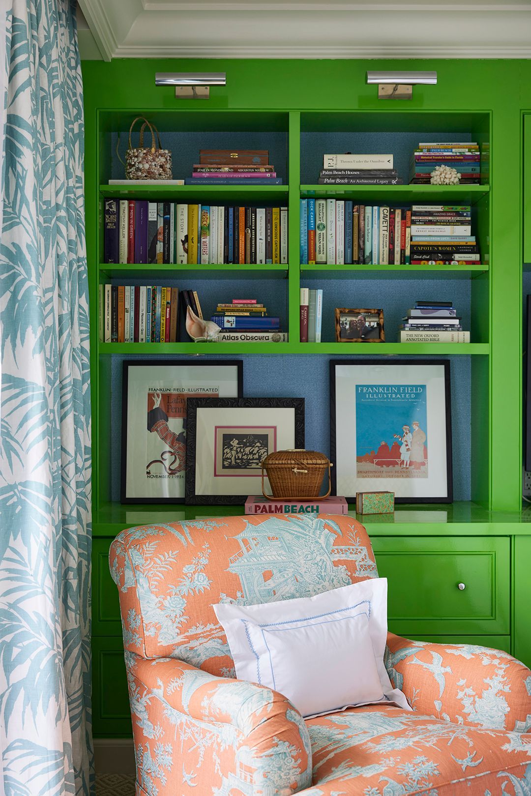 Green built-in bookshelves with books and art, against a blue wall. Orange patterned armchair with white pillows.