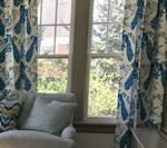 Two windows with blue patterned curtains frame a sunny outdoor view and a small reading nook.