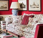 Floral couch with red trim in a red-walled room, lamp and framed art.