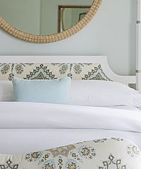 Bedroom with white bedding, patterned headboard, blue pillow, and woven mirror.