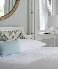 Bedroom with a bed, light blue pillows, patterned headboard, and a window with white shutters.