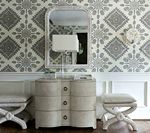 Bedroom with patterned wallpaper, mirror, chest of drawers, lamp, and two upholstered stools.