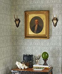 Corner of a room with portrait, mounted antlers, and decorative objects on a table, all against patterned wallpaper.