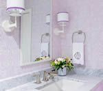 Bathroom with a white sink, mirror, and pink floral wallpaper. White sconces and towel ring with a monogrammed towel.