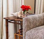 Side table with seashell and red flowers, beside patterned armchair and cream curtains.