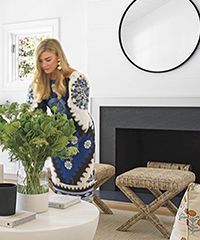 Woman arranging flowers in a modern living room with a fireplace and round mirror.