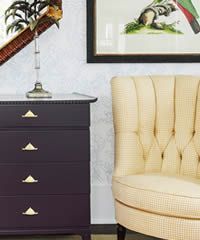 Dark purple dresser with gold drawer pulls, cream upholstered chair, artwork.