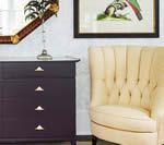 Dark purple dresser with gold drawer pulls, cream armchair, artwork, and mirror.