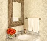 Bathroom with a rustic mirror, floral wallpaper, white towels, and a vase of orange flowers.