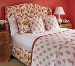 Floral upholstered bed in a pink bedroom; white bedding with pink spots, rattan nightstands, and lamp.