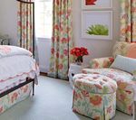 Bedroom with floral curtains, bedspread, armchair, and ottoman; white walls, green rug.