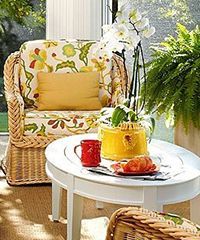Wicker chair with floral cushions, yellow pillow, white table with breakfast, and orchids, near a sunny window.