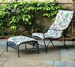 Black metal patio chair and ottoman with floral cushions; against a stone wall with green foliage.