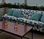 Outdoor seating area: floral sofa with blue cushions, dark wood coffee table with lit candles, greenery, and ocean view.