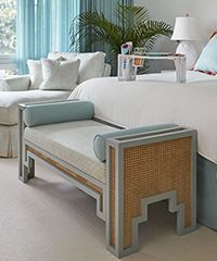 Bedroom with a white bed, chaise lounge, and a bench with cane detailing in a light-colored room with blue accents.