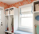 White built-in storage with window seat, orange patterned wall. Baskets, gear, and a window with a Roman shade.