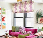 Brightly colored living room: pink sofa with patterned pillows, window with patterned shade, art on walls.