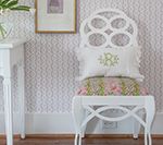 White chair with patterned back and seat, two pillows. Lilac wallpaper, wooden floor.