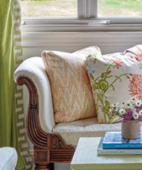 Couch with patterned pillows, wooden armrest, side table with books, window in background.