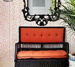 Black wicker bench with orange cushions against a pink speckled wall, ornate black mirror above.