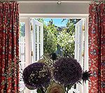 Open white French doors with a view of greenery, flanked by red patterned curtains. Flowers in foreground.
