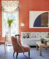 Living room with orange walls, floral chair, and a white sofa against a window with a patterned shade.