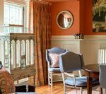 A dining room with orange walls, gray chairs, a round mirror, and a window with patterned curtains.