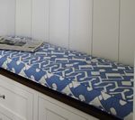 White built-in bench with blue patterned cushion, a newspaper on top. White cabinets below and panelled wall behind.