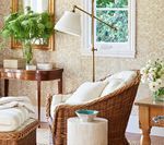 Wicker armchair with white cushions, lamp, side tables, window, and floral wallpaper.