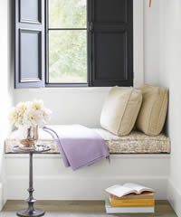 Cozy window seat with black shutters, beige cushions, lavender throw, and floral bouquet.