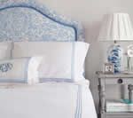 Bedroom with white and blue decor; upholstered headboard, bedding, lamp on gray nightstand.