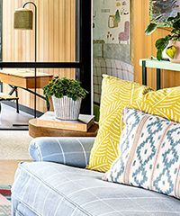 Couch with yellow and patterned pillows. A plant and a desk are in the background, by a wooden wall.