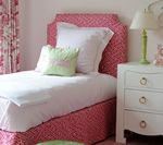 Pink patterned bed and headboard in a light-colored bedroom with white nightstand and green lamp.