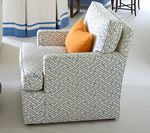 Upholstered armchair with geometric pattern, orange pillow, light blue and white curtains in background.