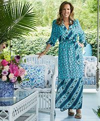 Woman in blue floral dress on porch with wicker furniture and plants.