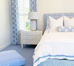 Bedroom with blue and white decor: bed, curtains, nightstand, floor pouf, and window.