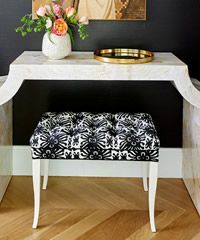 Marble-topped console table with a patterned black and white upholstered stool in front; flowers and a gold tray.