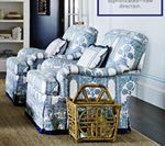 Two floral-patterned armchairs with pillows, near a wicker basket and a bookshelf.