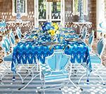 Outdoor dining table set for guests with blue patterned tablecloth, chairs, and centerpieces.