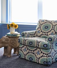 Floral armchair, wooden elephant side table with vase of sunflowers, against blue wall and window.