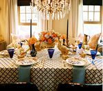 Elegant dining table set for a meal. Black patterned tablecloth, orange floral centerpiece, blue glasses.