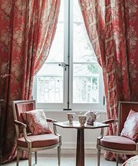 Red floral curtains frame a window overlooking a garden. Two armchairs and a small table with tea set are in front of the window.