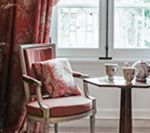 A small chair with a floral pillow and a side table with teacups sit by a window with red curtains.