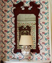 Mirror with ornate frame hanging on patterned wallpaper, reflecting a lamp.