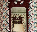 Dark wood framed mirror reflecting a chair and lamp against a floral patterned wallpaper.