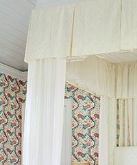 Cream-colored canopy bed with long white sheer curtains against patterned wallpaper.