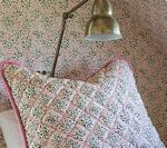 Floral patterned pillow with pink trim and a brass lamp against floral wallpaper.