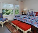 Bedroom with floral bedding, red ottomans, armchair, and a window overlooking greenery.