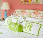 Bed with white linens, floral headboard, and monogrammed green pillows against a pink wall.