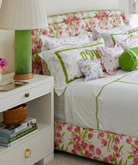 Nightstand beside a bed with floral headboard. Green lamp and white bedding with green trim.